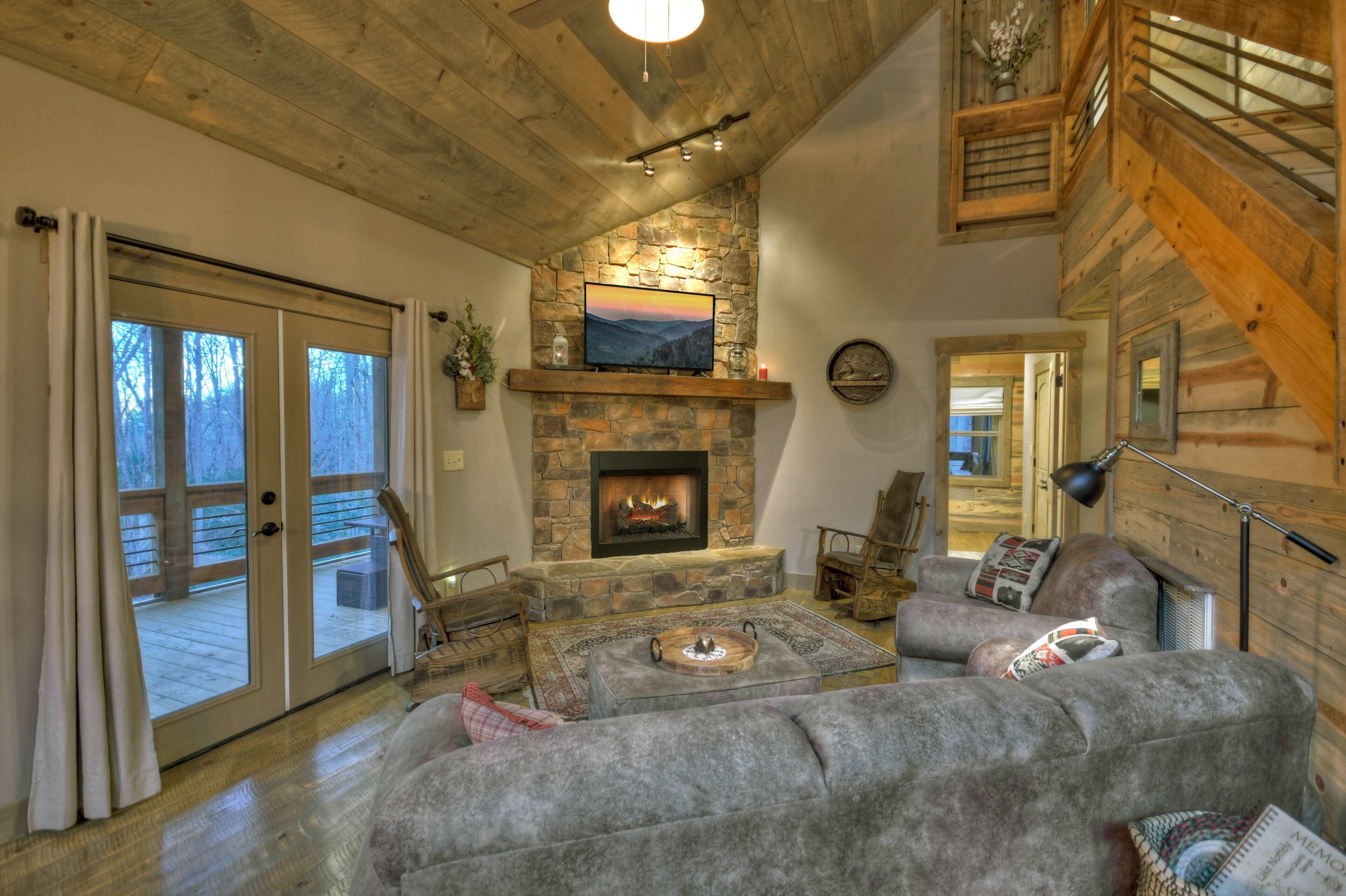 Un salon chaleureux avec une cheminée en pierre, un plafond en bois et une vue sur l’extérieur grâce à de grandes portes coulissantes.