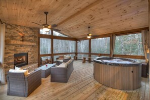 Una terraza de madera con chimenea de piedra, mobiliario de mimbre, un jacuzzi y grandes ventanales con vistas a un bosque.