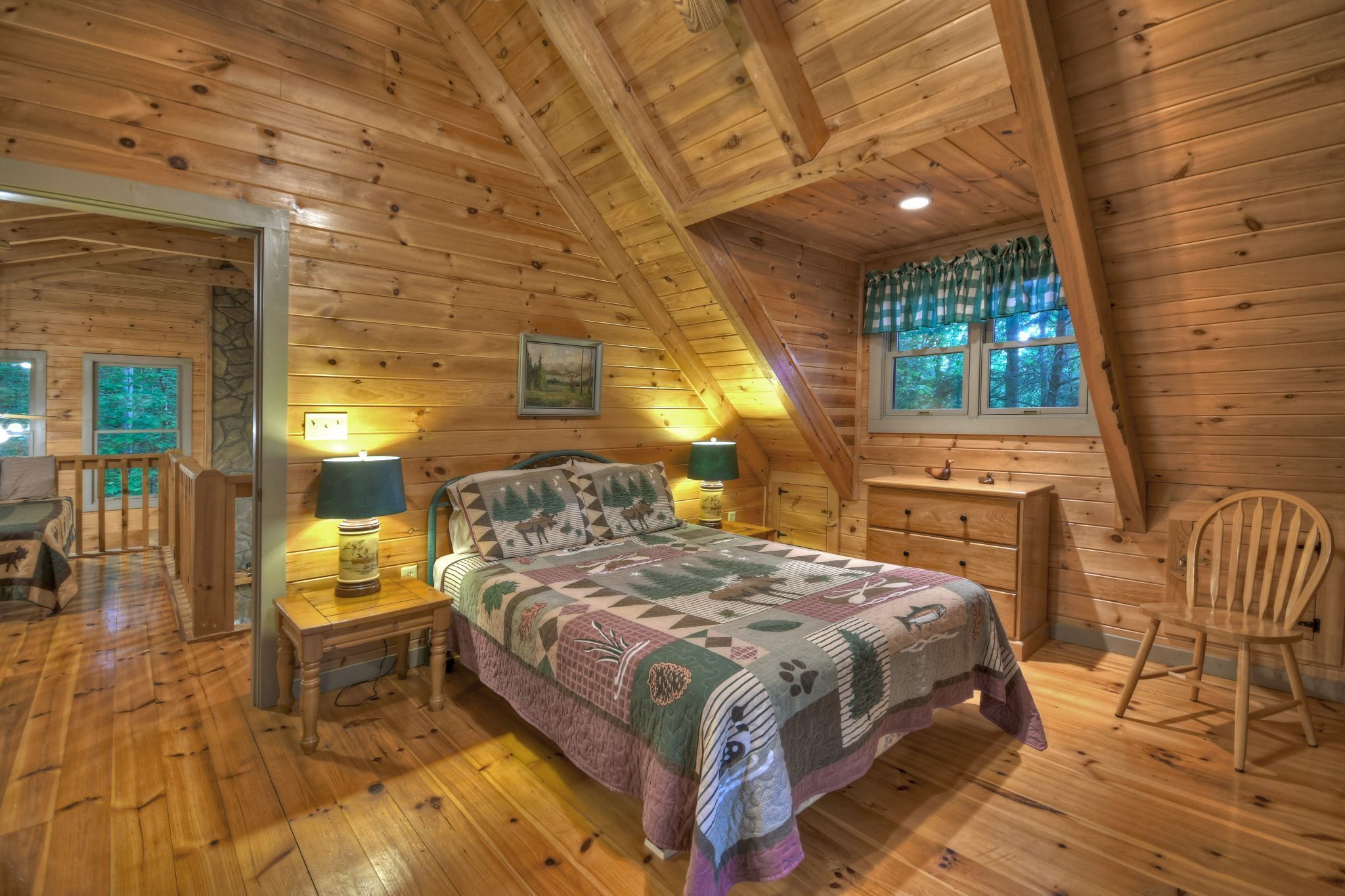A cozy bedroom with a wooden ceiling, a bed with a patterned quilt, two bedside lamps, and a window with a view of trees.