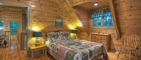 A cozy bedroom with a wooden ceiling, a bed with a patterned quilt, two bedside lamps, and a window with a view of trees.