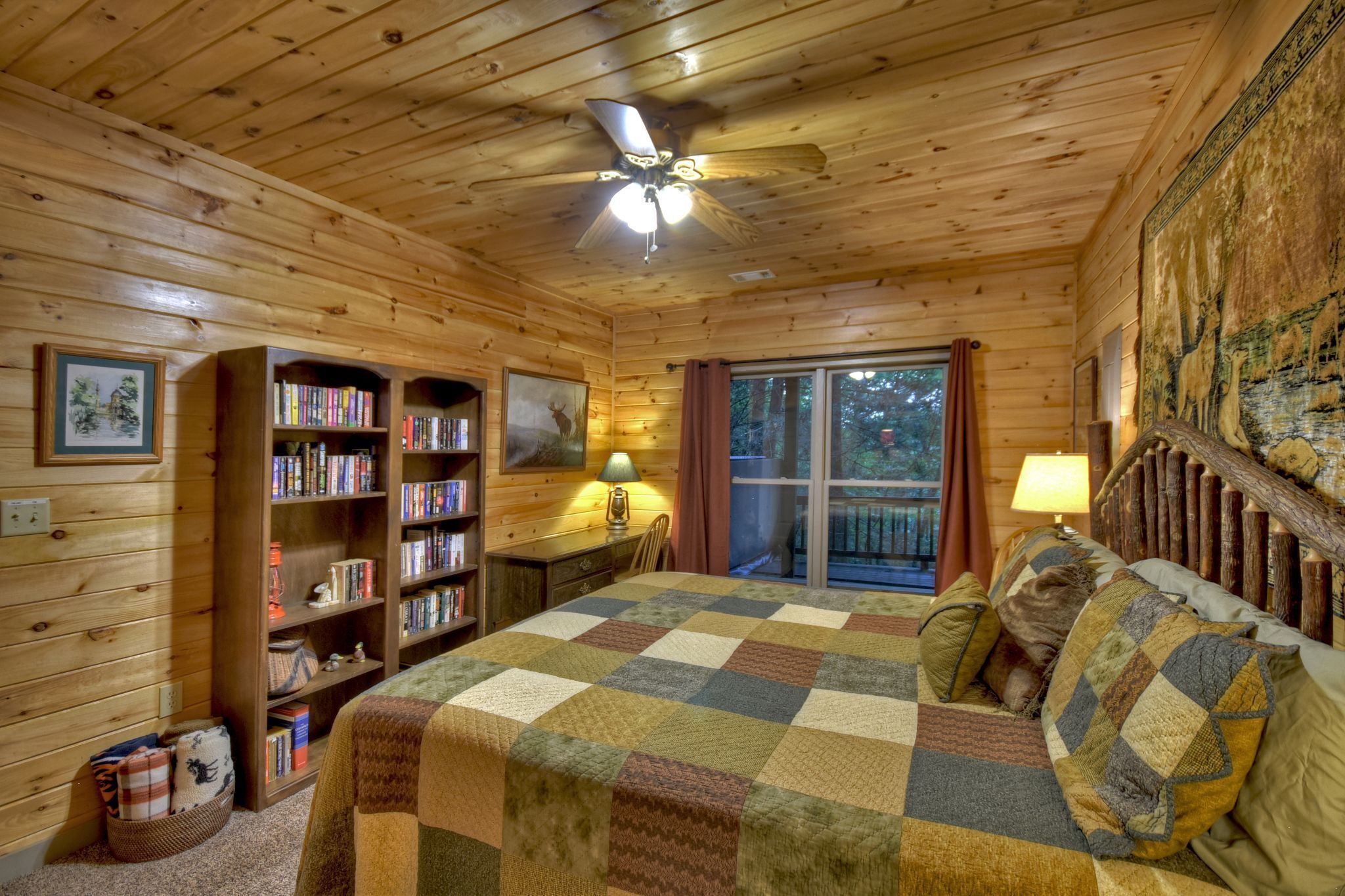 A cozy log cabin bedroom with a large bed, bookshelves, a desk, and a window with curtains.