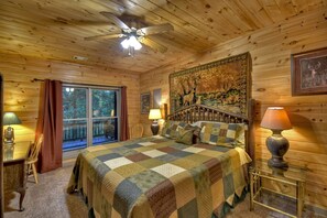 Une chambre de style cabane en bois, avec un grand lit, un ventilateur de plafond, une fenêtre donnant sur la forêt et une tapisserie au mur.