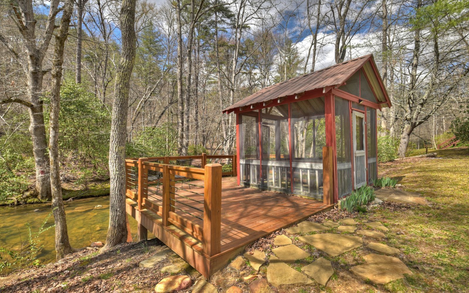 A wooden deck leading to a small pavilion with a red roof, surrounded by a forested area.