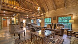 A spacious living area with a high ceiling, wooden beams, a fireplace, and large windows offering a view of the outdoors. - Knotty Creek Retreat - Creek access & hot tub (Morganton)