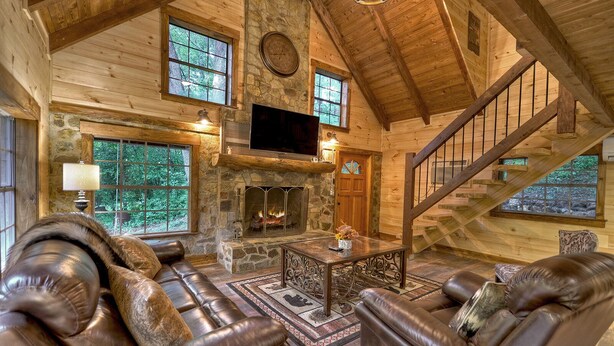 A log cabin interior with a stone fireplace, leather sofas, a wooden staircase, and large windows. - Knotty Creek Retreat - Creek access & hot tub (Morganton)