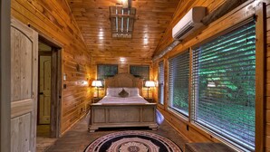 A wooden cabin bedroom with a bed, two bedside lamps, a circular rug, and a window with blinds. - Knotty Creek Retreat - Creek access & hot tub (Morganton)