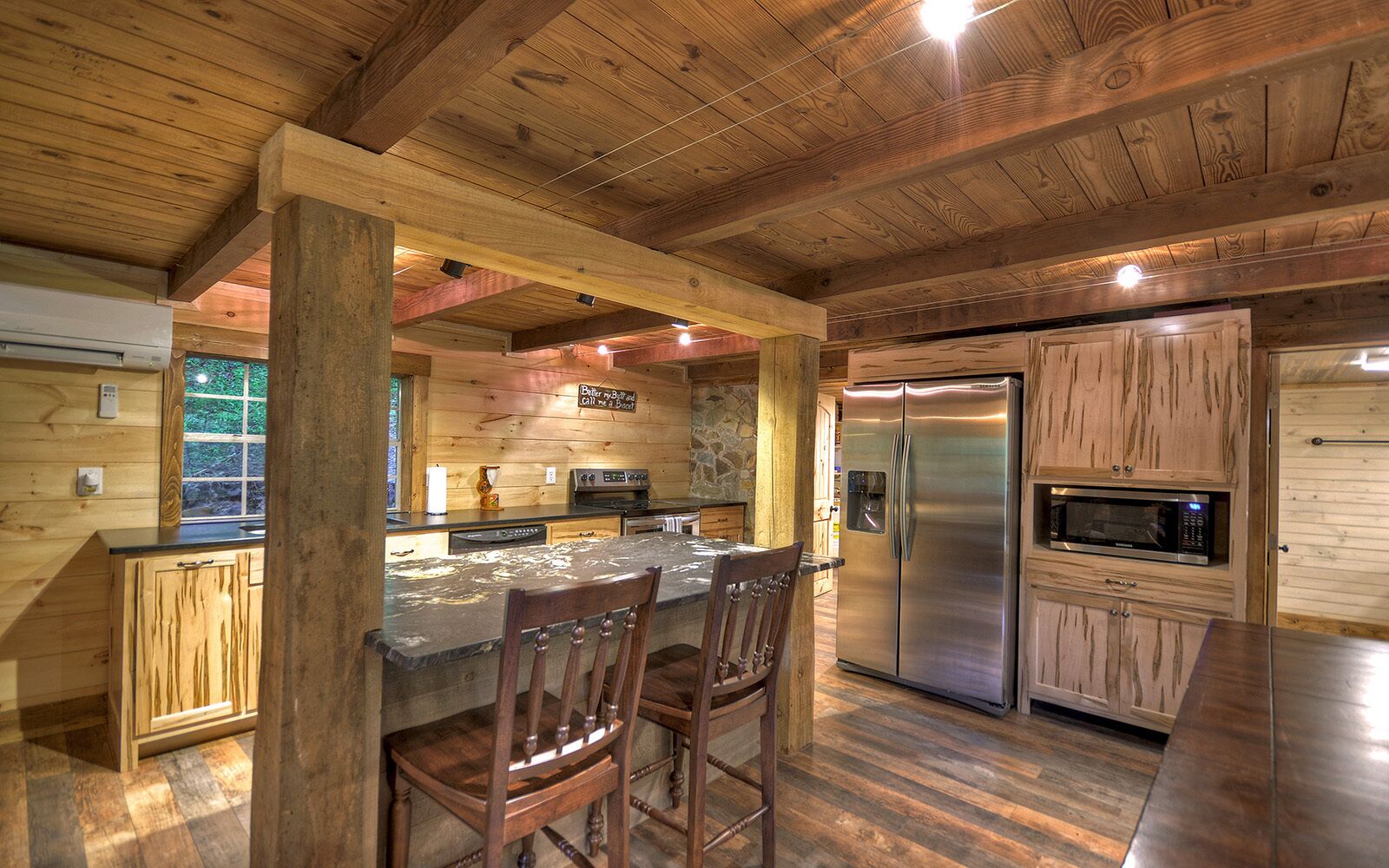 A rustic kitchen with wooden beams, a central island with bar stools, a stainless steel refrigerator, and a built-in microwave.