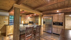 A rustic kitchen with wooden beams, a central island with bar stools, a stainless steel refrigerator, and a built-in microwave. - Knotty Creek Retreat - Creek access & hot tub (Morganton)