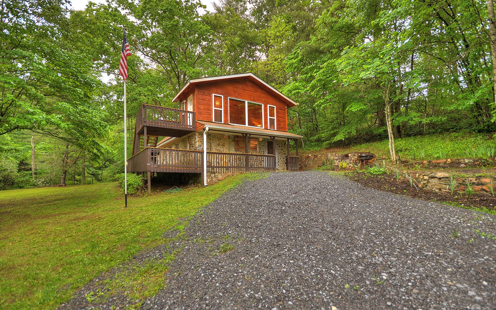 A rustic cabin with a porch, surrounded by lush greenery and a gravel driveway.