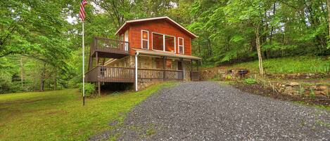 A rustic cabin with a porch, surrounded by lush greenery and a gravel driveway.