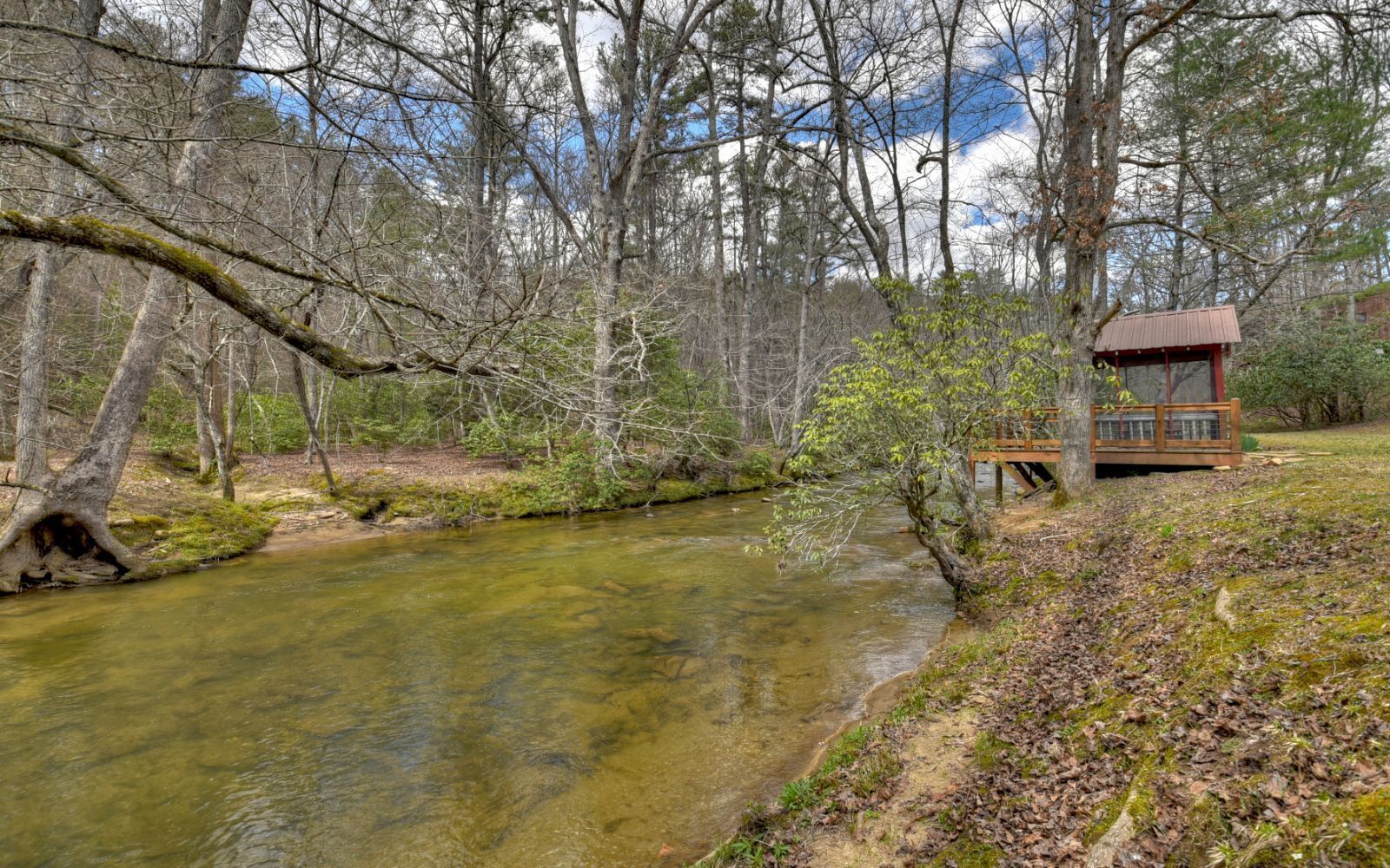 A wooden cabin by a clear stream surrounded by trees.