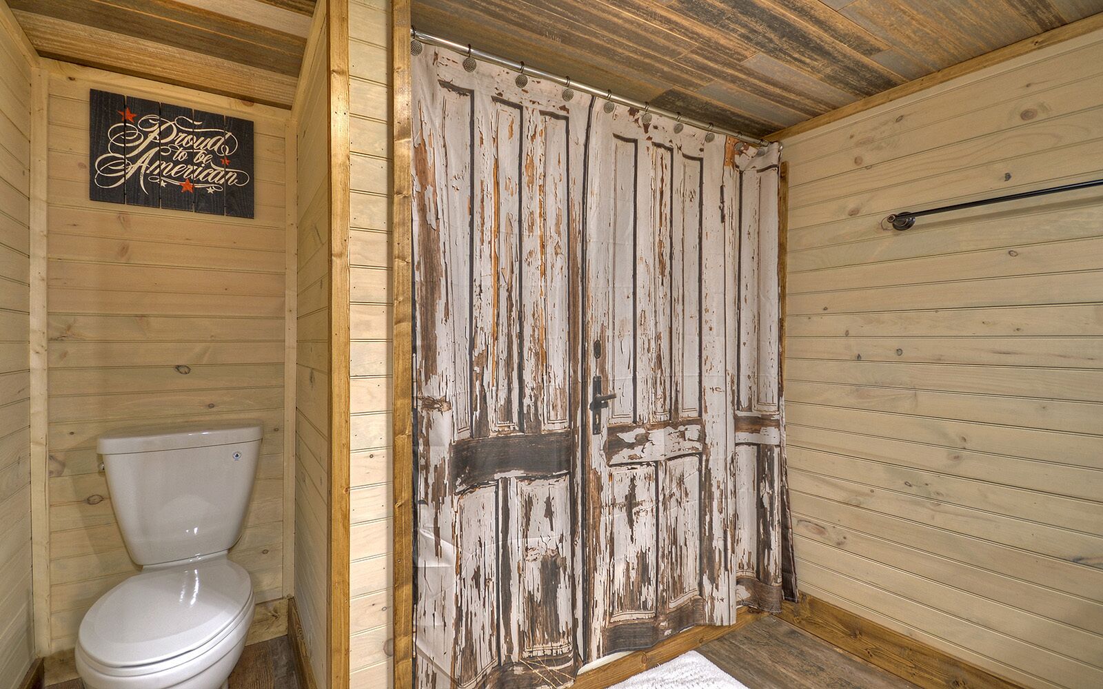 A rustic wooden bathroom with a white toilet, a weathered wooden door, and a 'Proud to be American' sign.