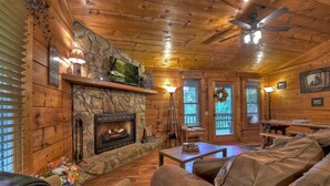 A cozy cabin interior with a stone fireplace, wooden walls, a ceiling fan, and a dining area. - Mama Bear's Hideaway - Hot tub, firepit, and more! (Morganton)