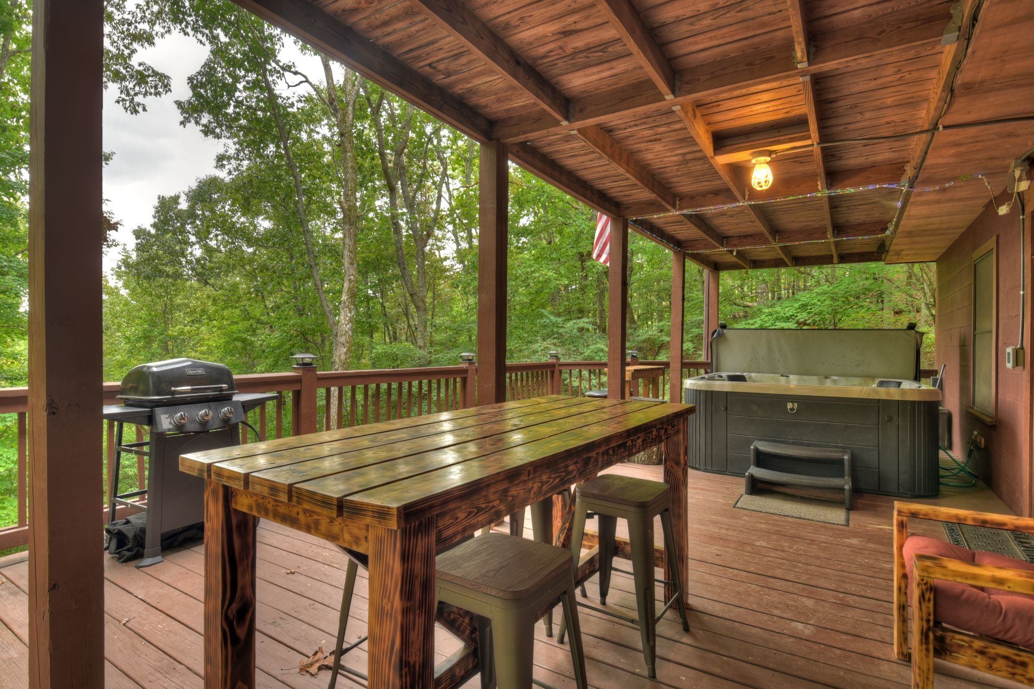 A covered outdoor deck with a wooden table, bar stools, a grill, and a hot tub.