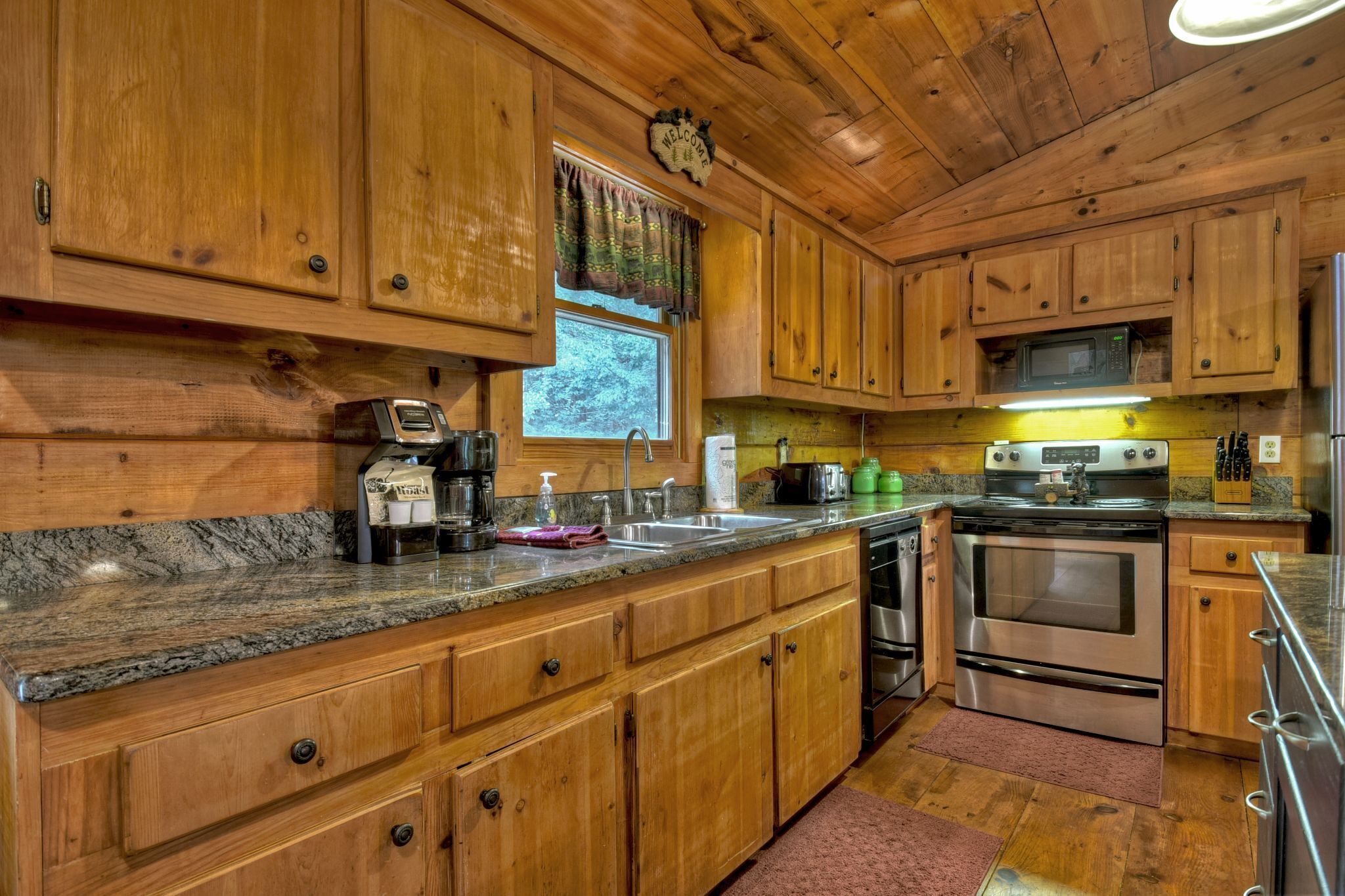 A wooden kitchen with granite countertops, stainless steel appliances, and a window with a curtain.