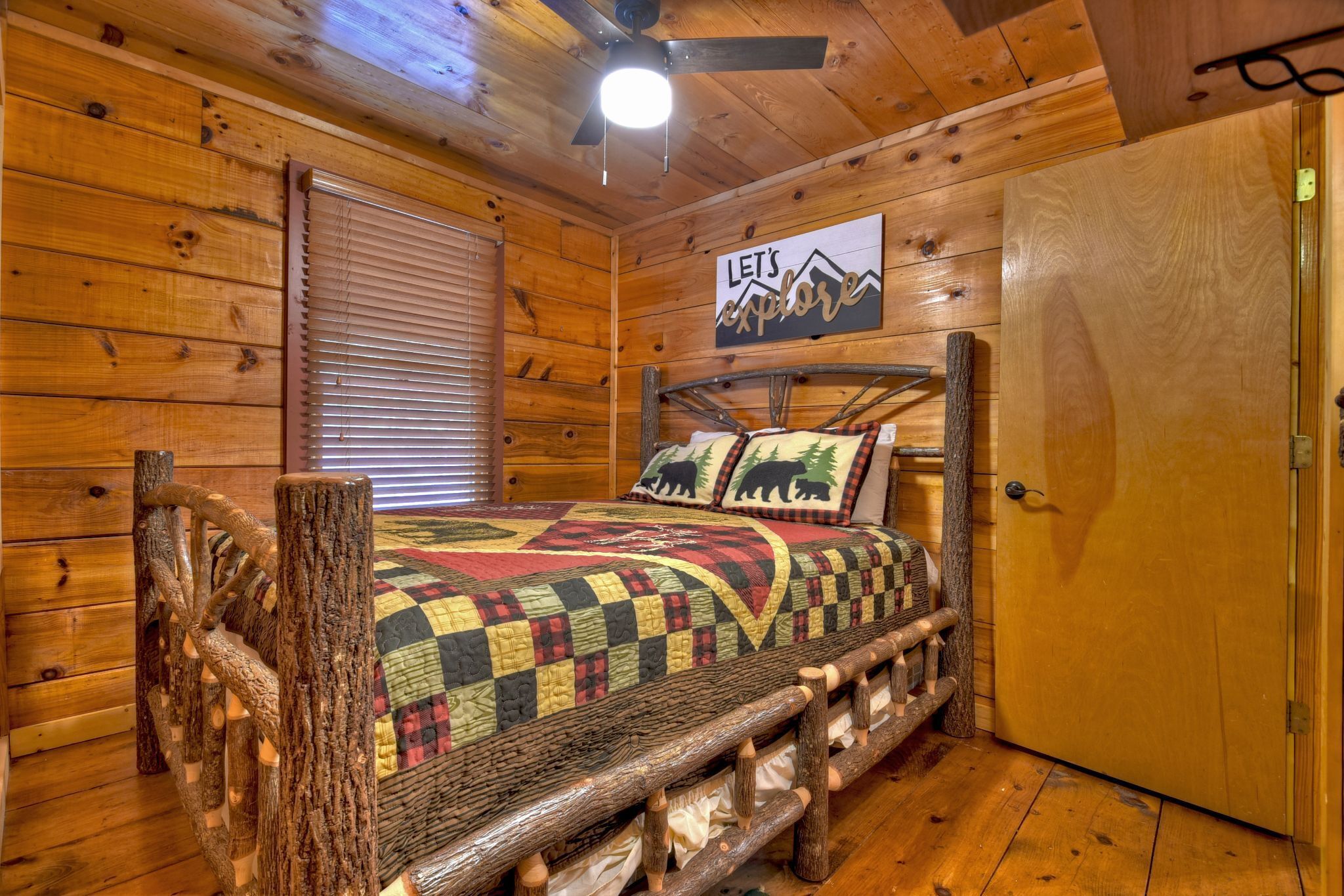 A wooden cabin room with a bed, a ceiling fan, a sign that says "LET'S EXPLORE," and a door.