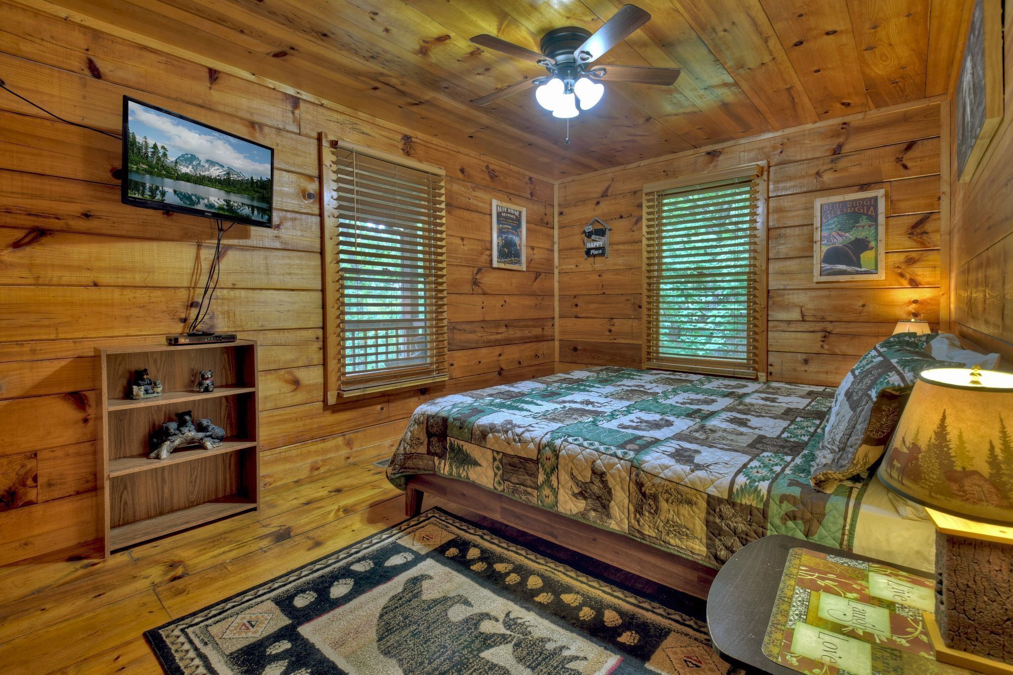 A wooden cabin bedroom with a bed, a television mounted on the wall, a ceiling fan, and a window with blinds.