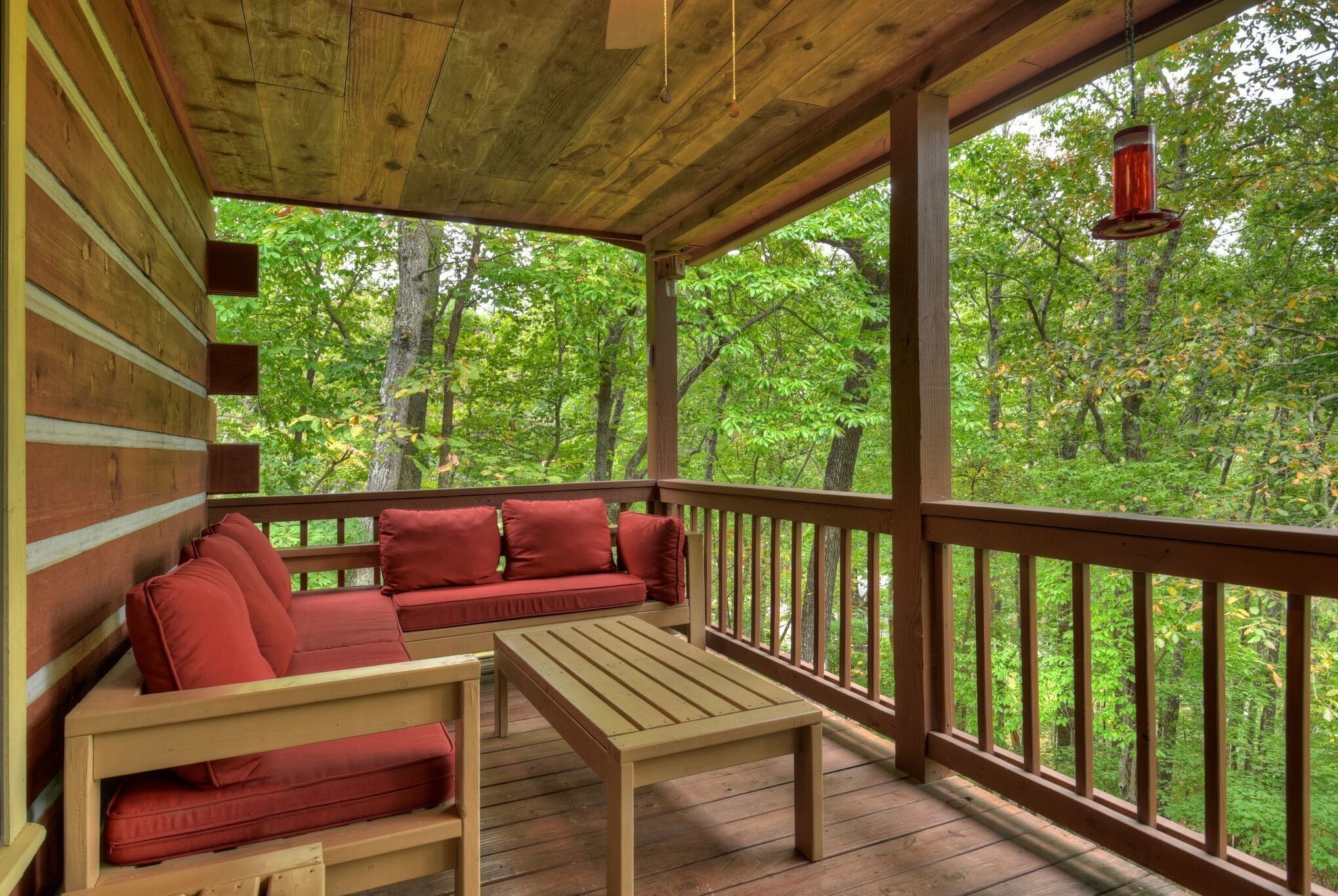 A wooden deck with a red sofa and a table, surrounded by a railing with a view of a forest.