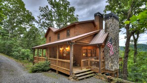 A log cabin with a stone chimney, wooden porch, and an American flag.