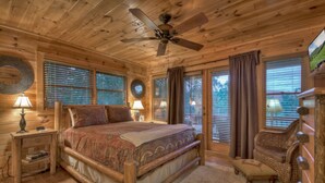 A wooden cabin bedroom with a large bed, two bedside tables with lamps, a chair, and a ceiling fan.