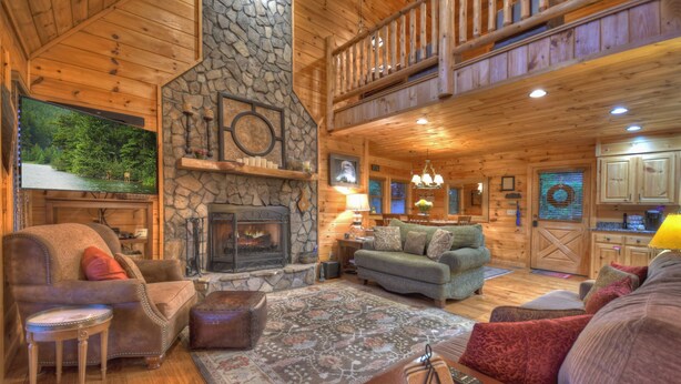 A cozy cabin interior with a stone fireplace, wooden floors, a balcony, and a large window overlooking a forest.