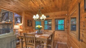 A wooden cabin interior with a dining table, chairs, a chandelier, a fireplace, and a couch.