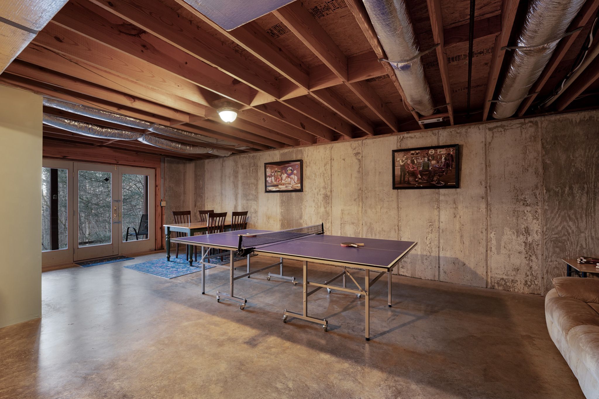 Une table de ping-pong dans une pièce aux poutres en bois et aux murs en béton, une porte donnant sur l’extérieur et deux tableaux encadrés accrochés au mur.
