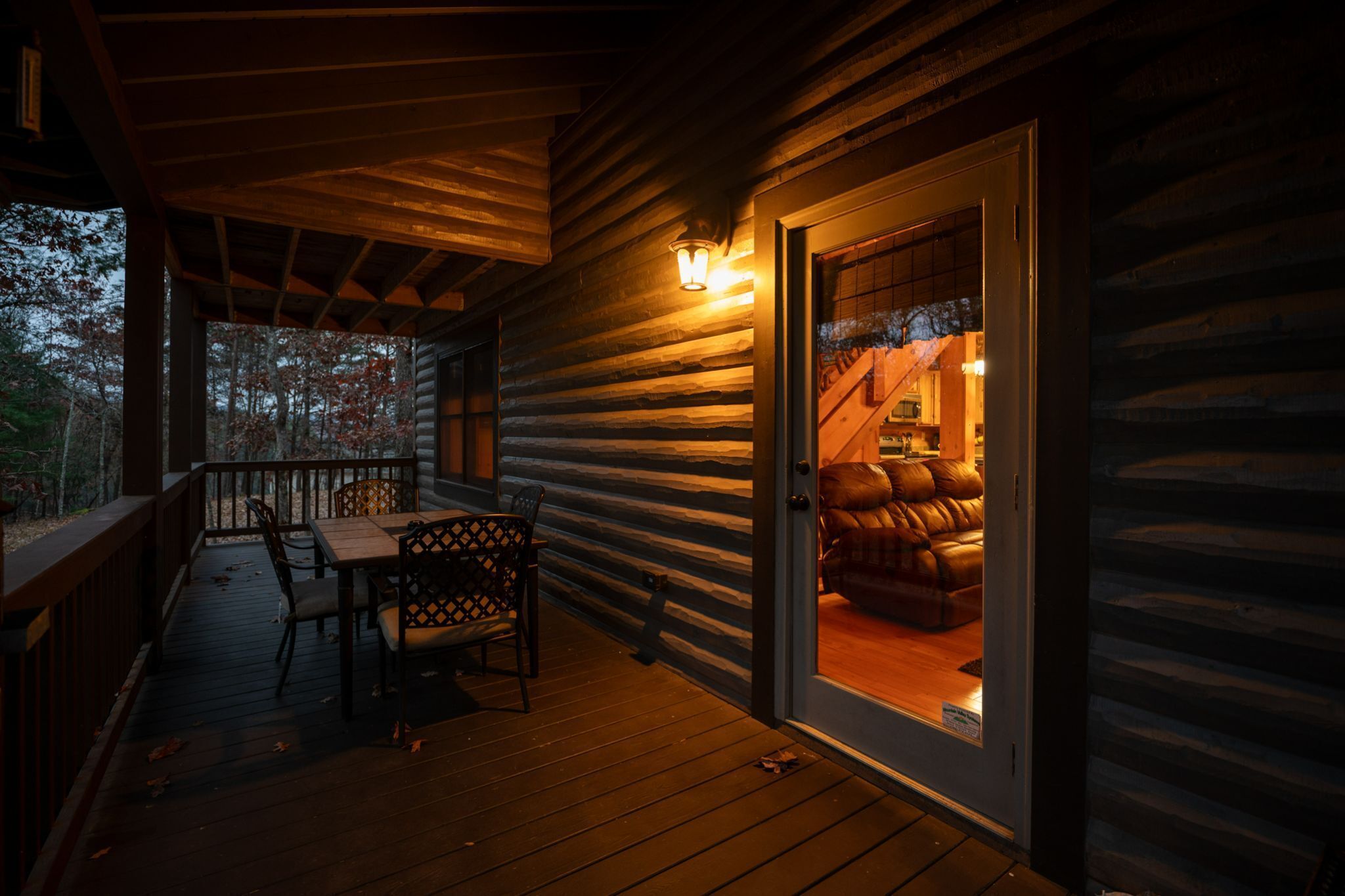 A log cabin with a wooden deck, a sliding glass door leading to a cozy interior with a leather sofa and a table.