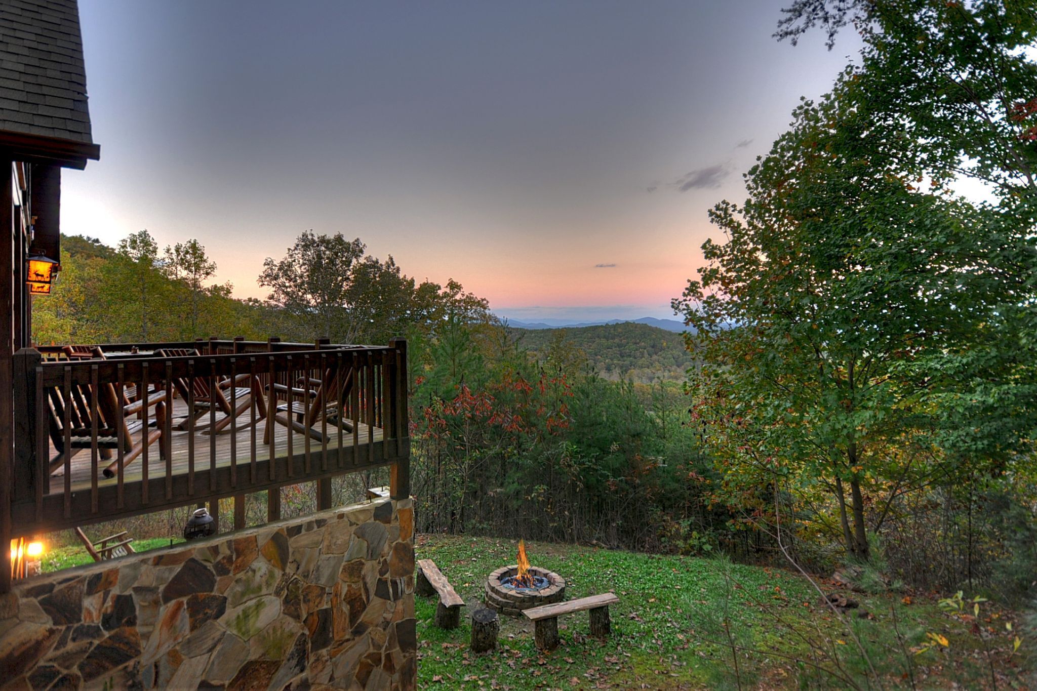 A stone building with a wooden deck and chairs, a fire pit, and a view of the surrounding forest and mountains.