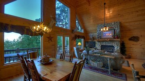 Interior de una cabaña de madera con comedor, chimenea y ventanas amplias.