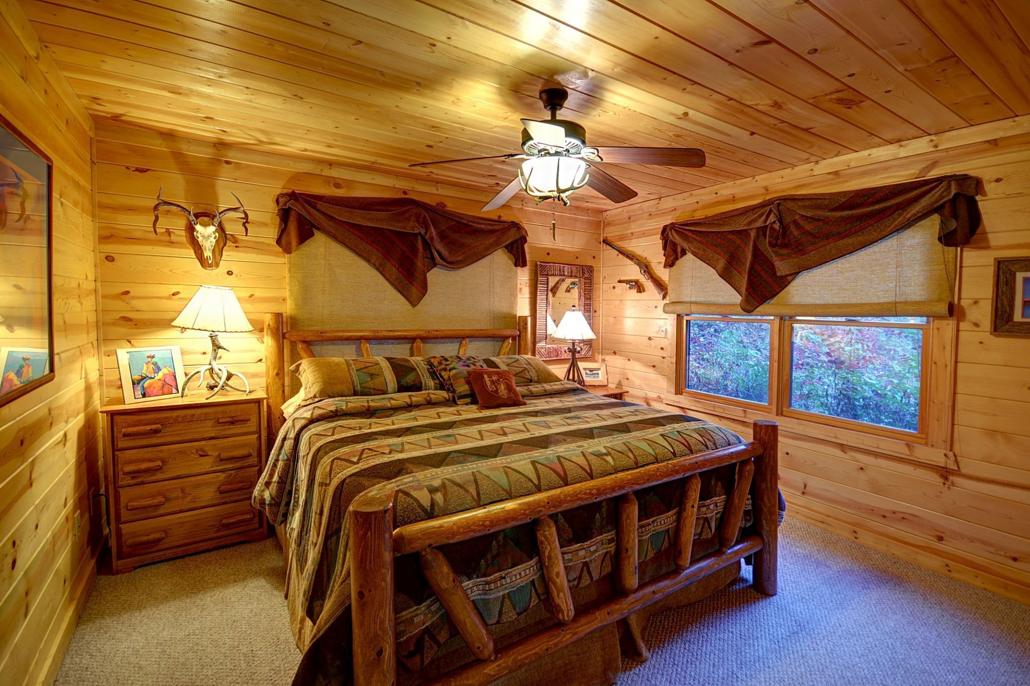 Un dormitorio de cabaña de madera con una cama, dos lámparas de noche, un ventilador de techo y ventanas con vistas a árboles.