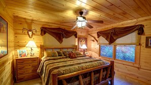 Un dormitorio de cabaña de madera con una cama, dos lámparas de noche, un ventilador de techo y ventanas con vistas a árboles.