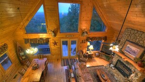 Interior acogedor de una cabaña con mesa de comedor de madera, sofá de cuero, chimenea de piedra y ventanas amplias con vistas al bosque.