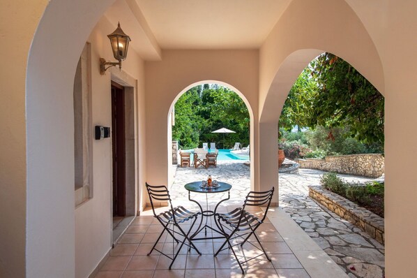 A covered outdoor patio with a table and chairs, a pool, and a stone wall.
