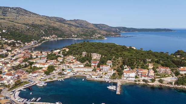 Eine Küstenstadt mit einer Marina, Gebäuden mit roten Dächern und einem Hügel voller üppiger Vegetation.