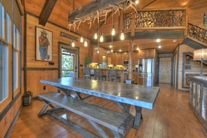 A rustic cabin interior with a large wooden dining table, a kitchen area, and a staircase leading to a loft. - Skyfall - Stunning mountain views, cozy hot tub, comfy beds, game room, and more! (Mineral Bluff)