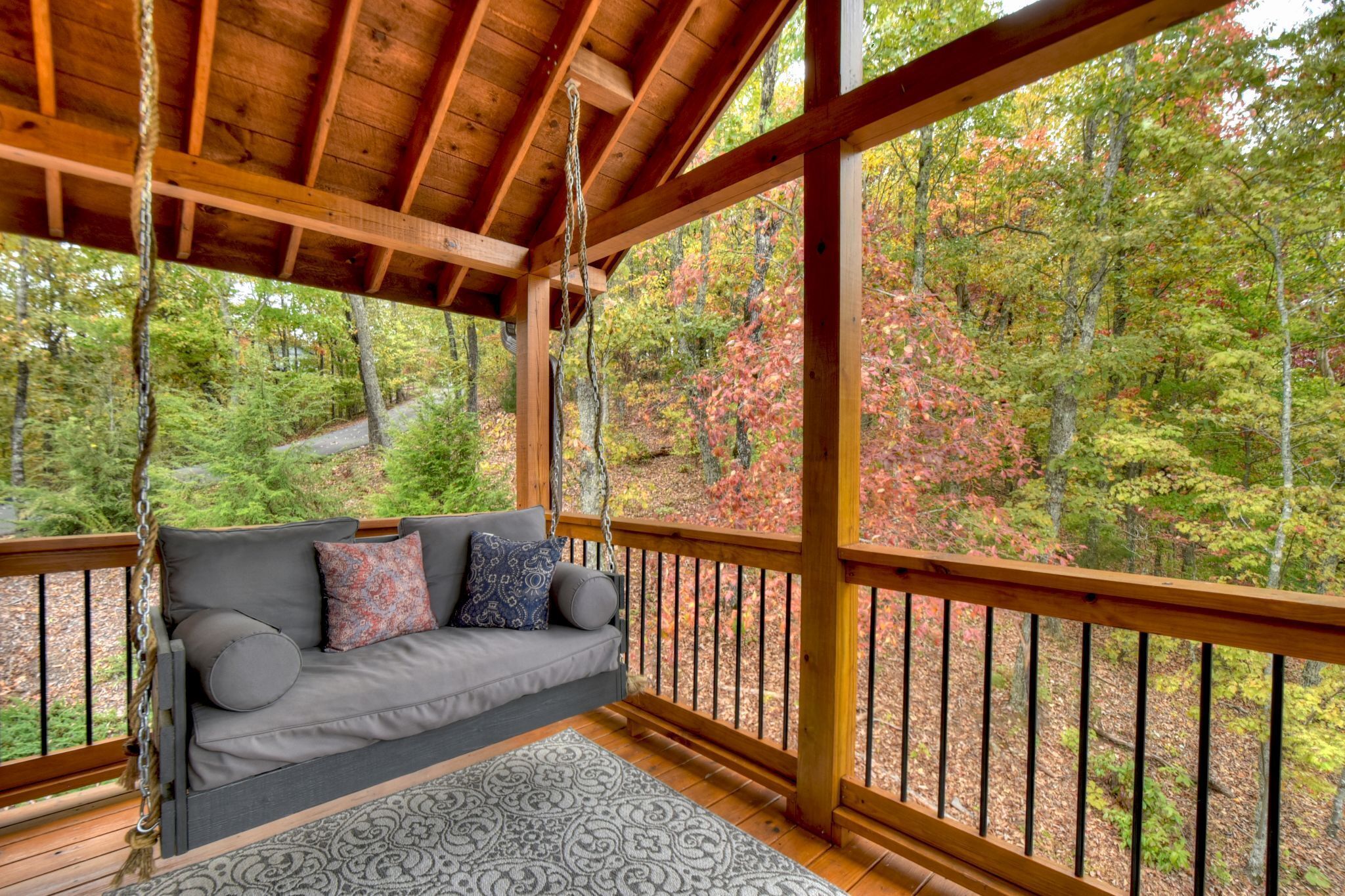 A wooden deck with a swing chair and a sofa facing a forested area.