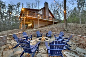 A cabin with a fire pit and Adirondack chairs, surrounded by a stone patio and a wooden deck. - Cozy Retreat with a Firepit & Fireplace- S'mores Cabin (Mineral Bluff)
