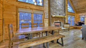 Interior de una cabaña de madera con una amplia mesa de comedor, bancos, una chimenea de piedra y ventanas grandes.