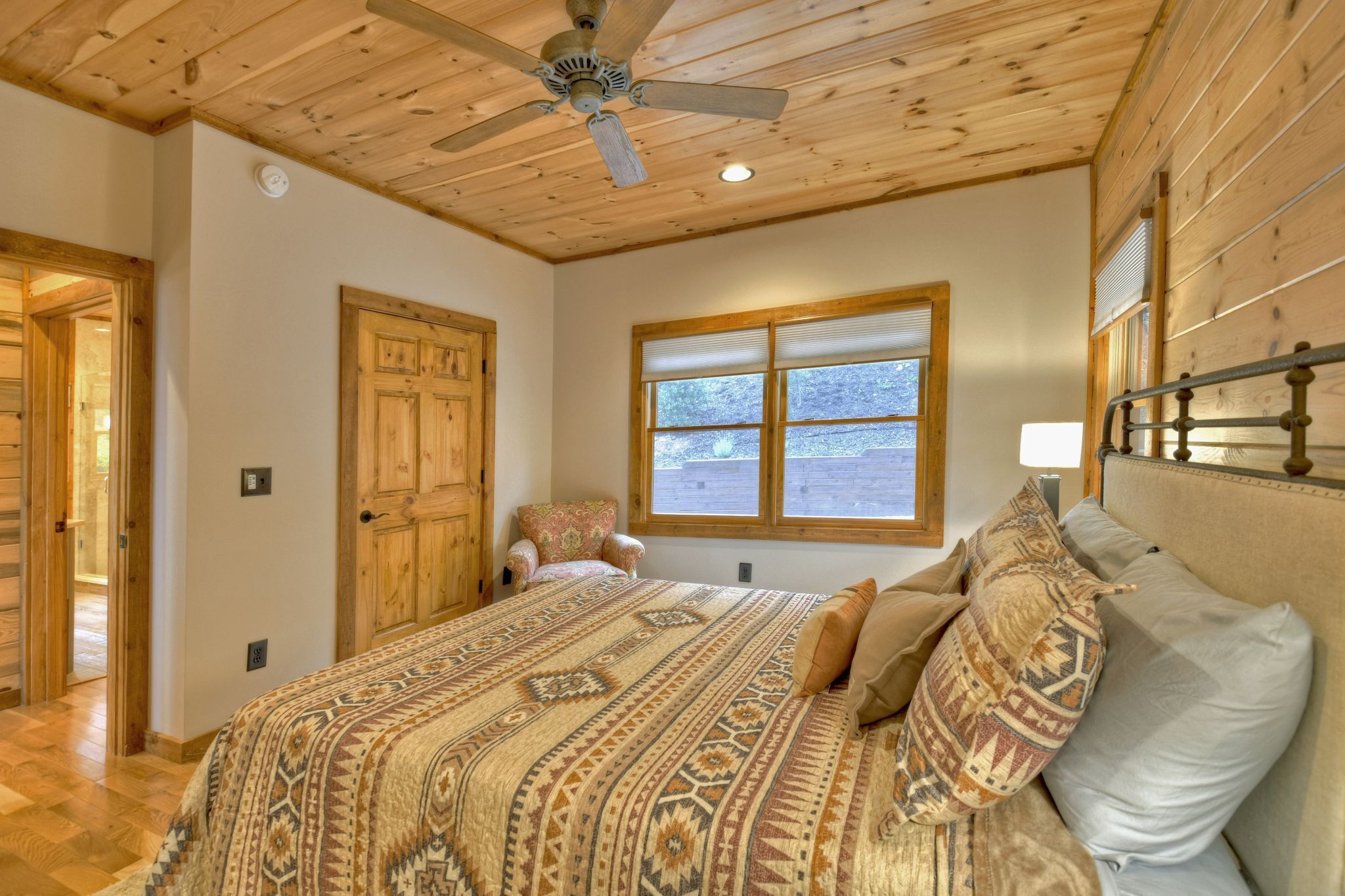 A bedroom with a wooden ceiling, a bed with patterned bedding, a chair, a window with a view, and a ceiling fan.