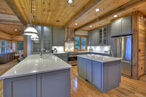 A modern kitchen with wooden ceiling beams, grey cabinetry, a large island, stainless steel appliances, and a view of the outdoors.