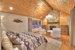 A cozy log cabin bedroom with a large bed, two white armchairs, a fireplace, and a stone wall.