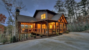 A large, two-story log cabin with a wrap-around porch, stone foundation, and a view of the surrounding forest. - Relax in private rustic luxury! - A Starry Night (Cherry Log)