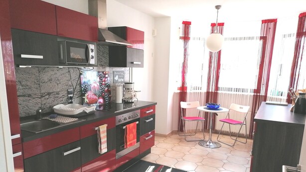 A modern kitchen with a microwave, coffee maker, and a sink. There are two chairs and a small table near a window with red curtains. - Beautiful 2-room granny apartment with terrace for 4 people (Burladingen)
