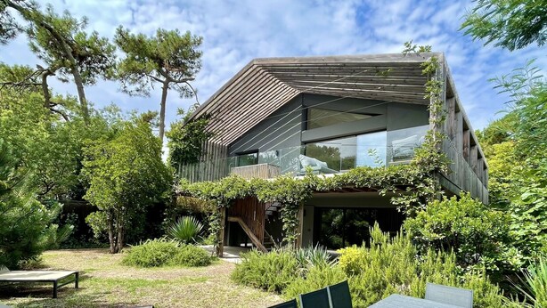 A modern house with a glass facade, surrounded by lush greenery and a patio area with outdoor furniture.