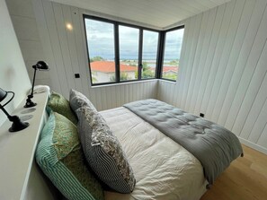 Een slaapkamer met een groot raam, een bed met veel kussens, een bedlampje en uitzicht op een gebouw met rode dakpannen.