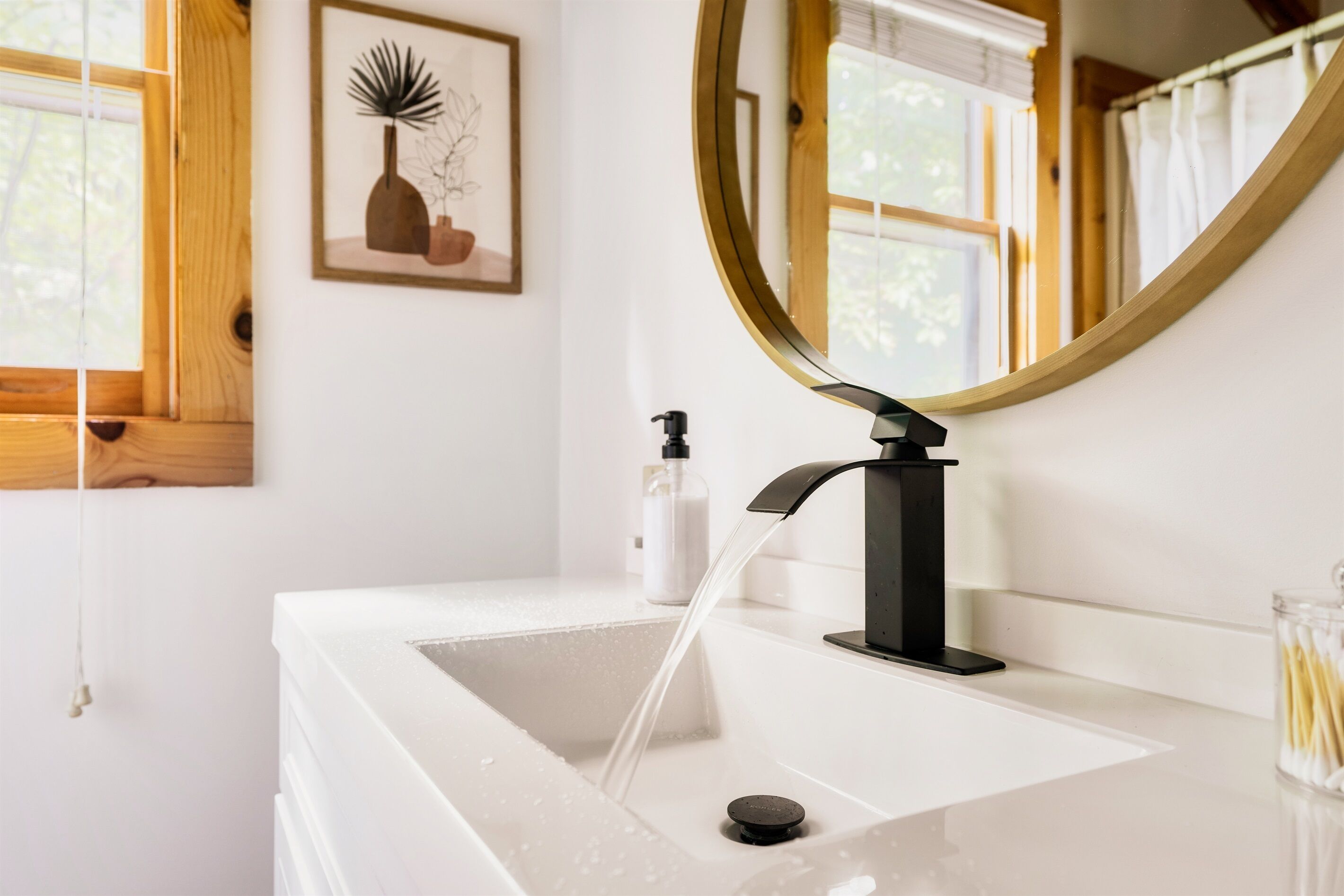 Un baño con un lavabo blanco, un grifo negro, un espejo redondo y una obra de arte enmarcada en la pared.