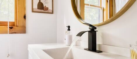 Un baño con un lavabo blanco, un grifo negro, un espejo redondo y una obra de arte enmarcada en la pared.