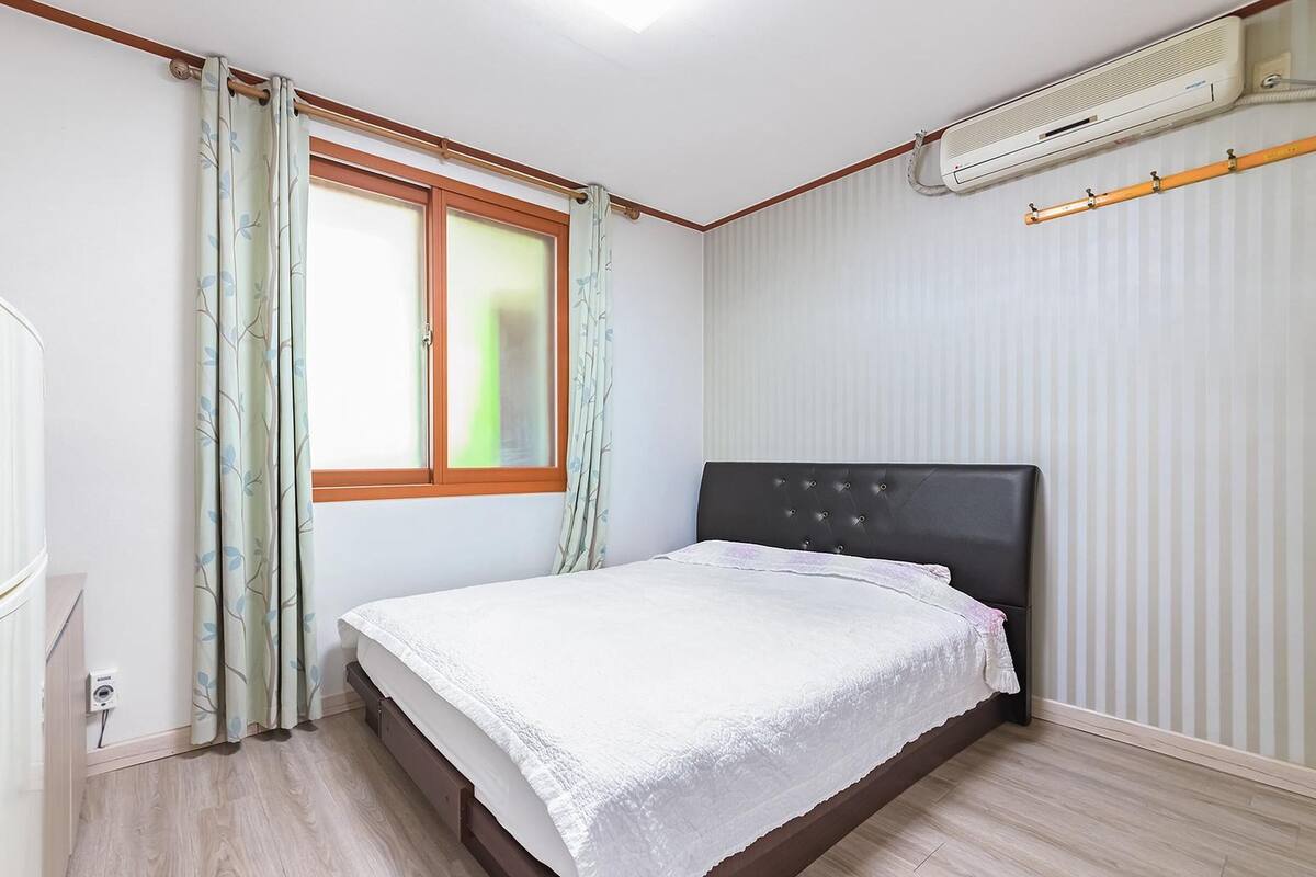 Une chambre à coucher avec un lit, une fenêtre avec des rideaux, un climatiseur et un plancher en bois.