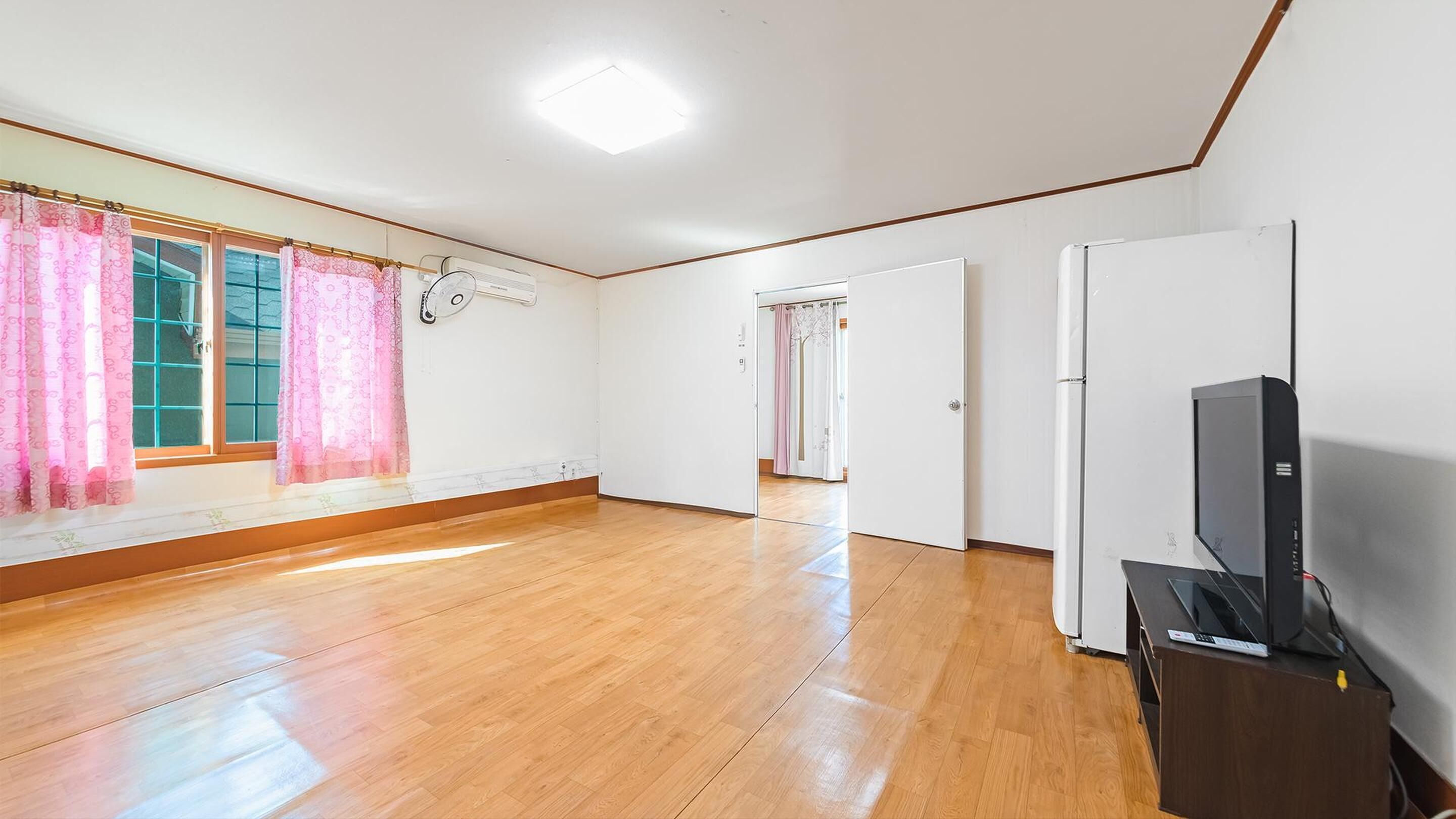 Una habitación amplia con piso de madera, un ventanal con cortinas rosadas, una puerta blanca y un televisor sobre un mueble de madera.